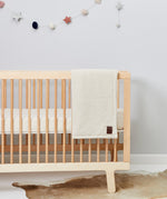 A linen coloured Little Green Sheep Organic Knitted Fleece Baby Blanket draped over the side of a baby cot