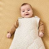 A baby wearing a linen rice print baby sleeping bag. The baby is lyinh down on a honey rice coloured cot sheet 