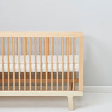 A cot pictured in a room in front of a light grey coloured wall. A Little Green Sheep Organic Cot Mattress Protector can be seen placed on the mattress inside the cot 