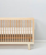 A cot pictured in a room in front of a light grey coloured wall. A Little Green Sheep Organic Cot Mattress Protector can be seen placed on the mattress inside the cot 