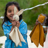 A child holding the Little Green Radicals rabbit organic cotton baby comforter toy in front of them. Another child is standing just to the side with a lion comforter. 