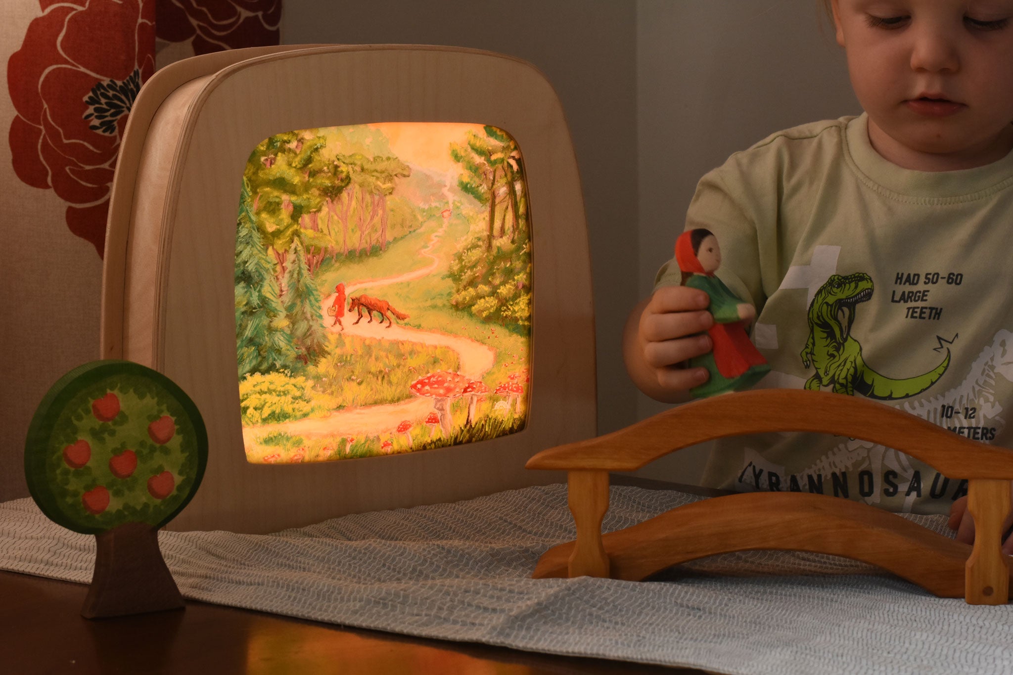 A Toverlux MagicLamp placed on a table with little red riding hood artwork placed inside. The lamp is on. There is a young child playing with an ostheimer little red riding hood wooden toy figure besides the lamp. 