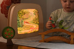 A Toverlux MagicLamp placed on a table with little red riding hood artwork placed inside. The lamp is on. There is a young child playing with an ostheimer little red riding hood wooden toy figure besides the lamp. 