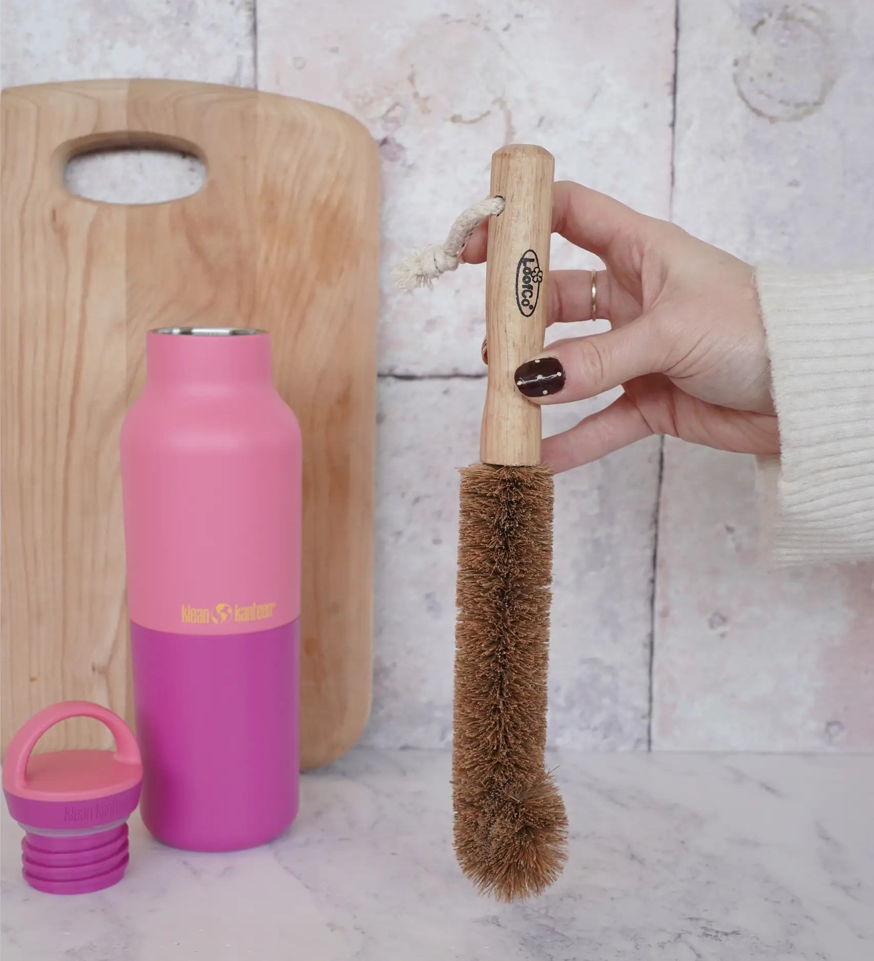 Adult hand holding a LoofCo coconut bottle brush with a Klean Kanteen pink bottle in the background
