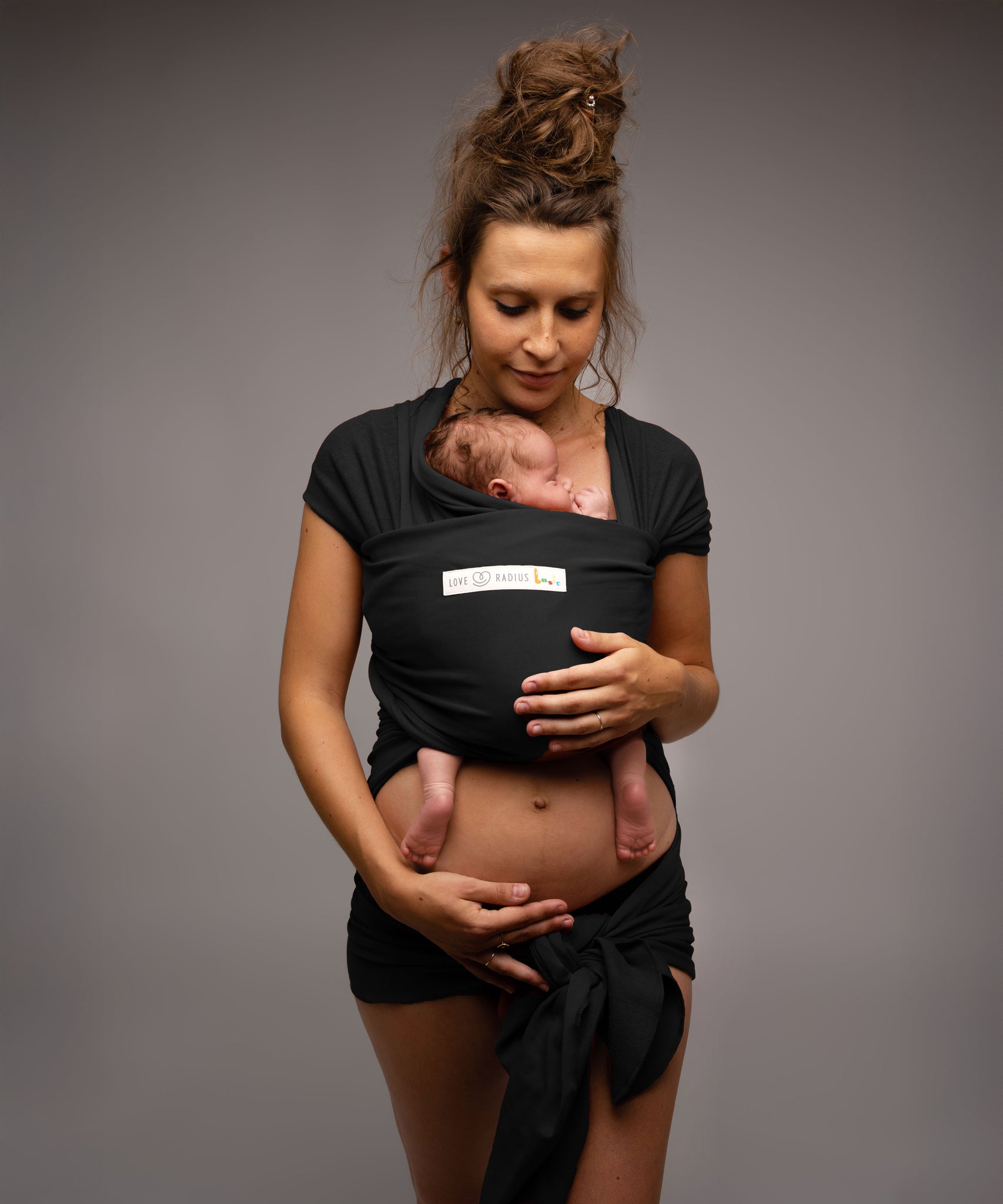 A mother wearing her baby in a black coloured Love Radius Basic Organic Baby Wrap. Babipur stock a wide range of organic baby wraps and carriers. 