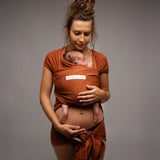 A mother wearing her baby in a fawn dark orange coloured Love Radius Basic Organic Baby Wrap. Babipur stock a wide range of organic baby wraps and carriers. 