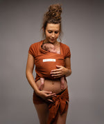 A mother wearing her baby in a fawn dark orange coloured Love Radius Basic Organic Baby Wrap. Babipur stock a wide range of organic baby wraps and carriers. 