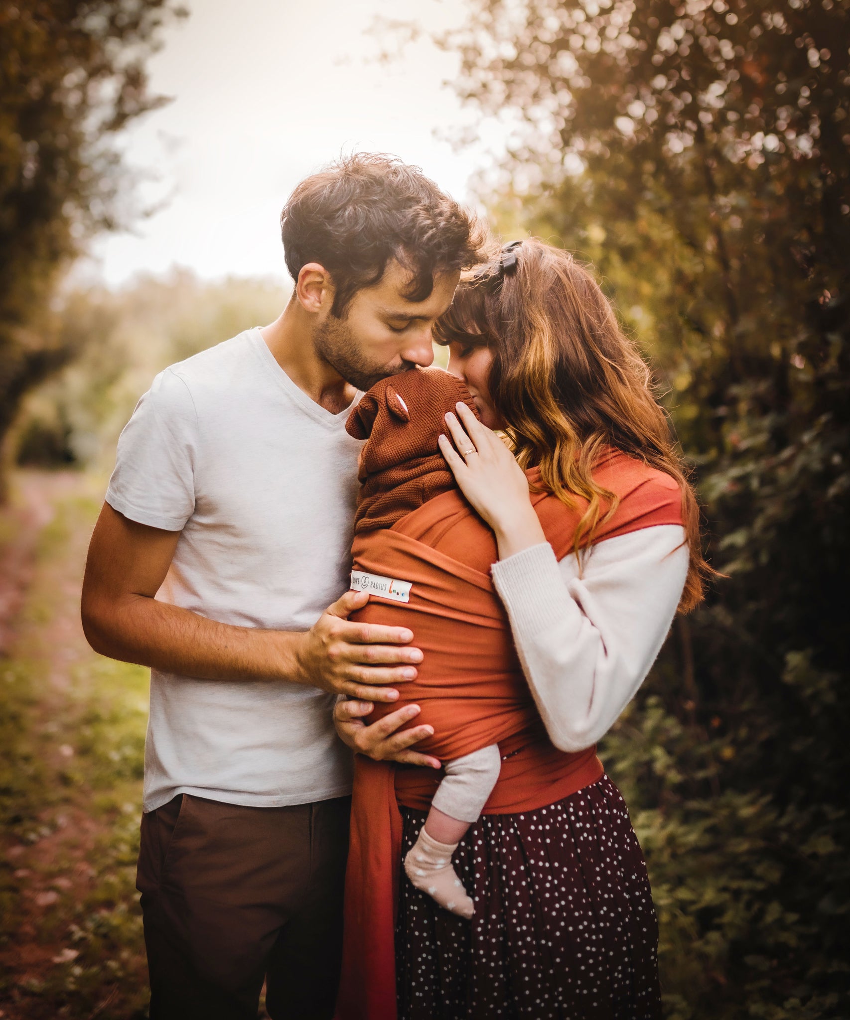 Two parents standing next to each other. The mother is wearing her baby in a fawn coloured Love Radius Basic Organic Baby Wrap. Babipur stock a wide range of organic baby wraps and carriers in various colurs. 