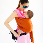 A parent wearing their baby in a fawn orange coloured Love Radius No Knot Woven Wrap. The parent is looking down at their baby with a smile of their face. A wide range of baby wraps and carriers are available at Babipur. 