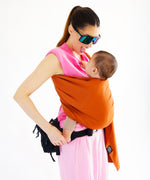A parent wearing their baby in a fawn orange coloured Love Radius No Knot Woven Wrap. The parent is looking down at their baby with a smile of their face. A wide range of baby wraps and carriers are available at Babipur. 