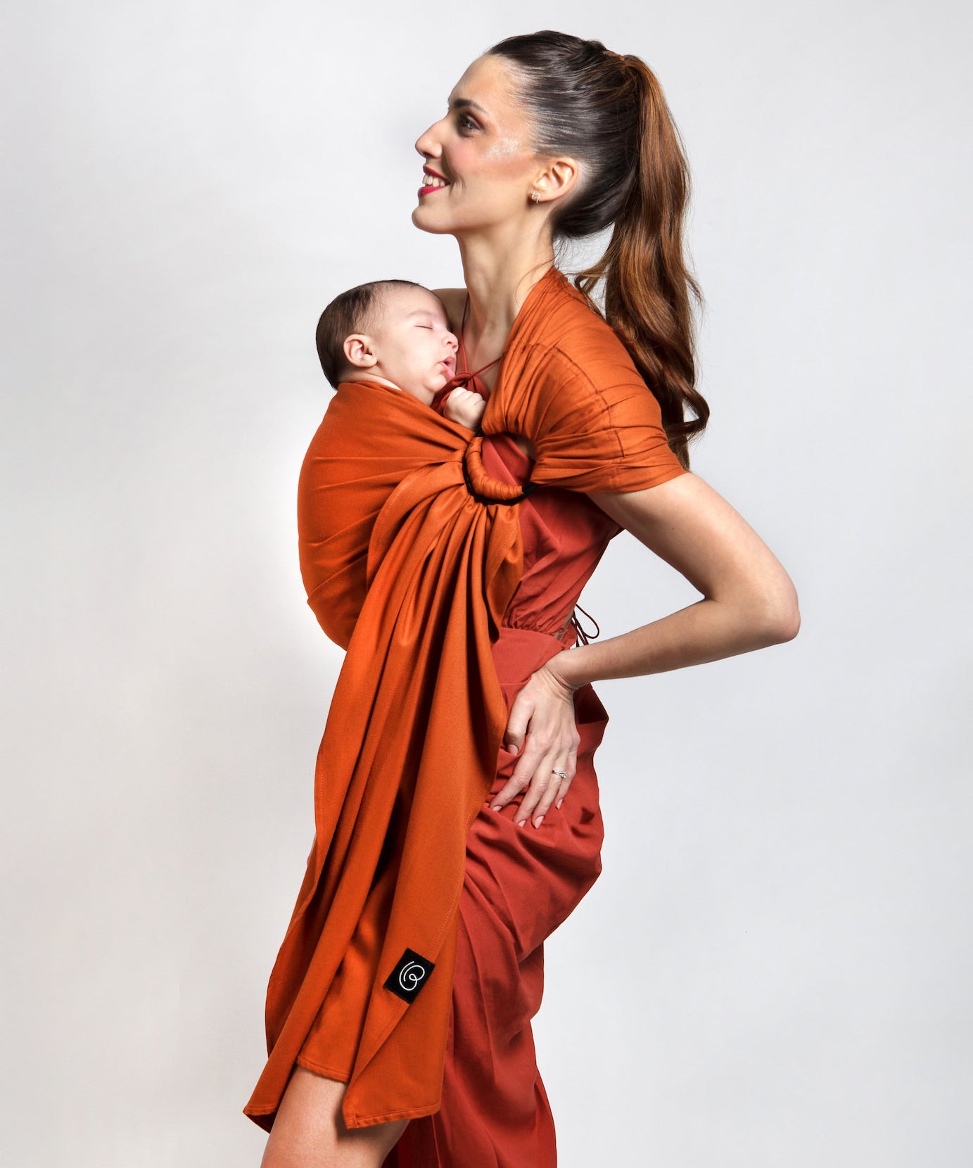 A parent wearing their young baby in a fawn orange coloured Love Radius No Knot Woven Wrap. A wide range of baby wraps and carriers are available at Babipur. 