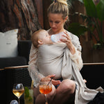 A parent wearing their baby in a white gold coloured Love Radius No Knot Woven Wrap. A wide range of baby wraps and carriers are available at Babipur. The parent is bottle feeding their baby whilst sitting down outdoors.