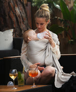 A parent wearing their baby in a white gold coloured Love Radius No Knot Woven Wrap. A wide range of baby wraps and carriers are available at Babipur. The parent is bottle feeding their baby whilst sitting down outdoors.