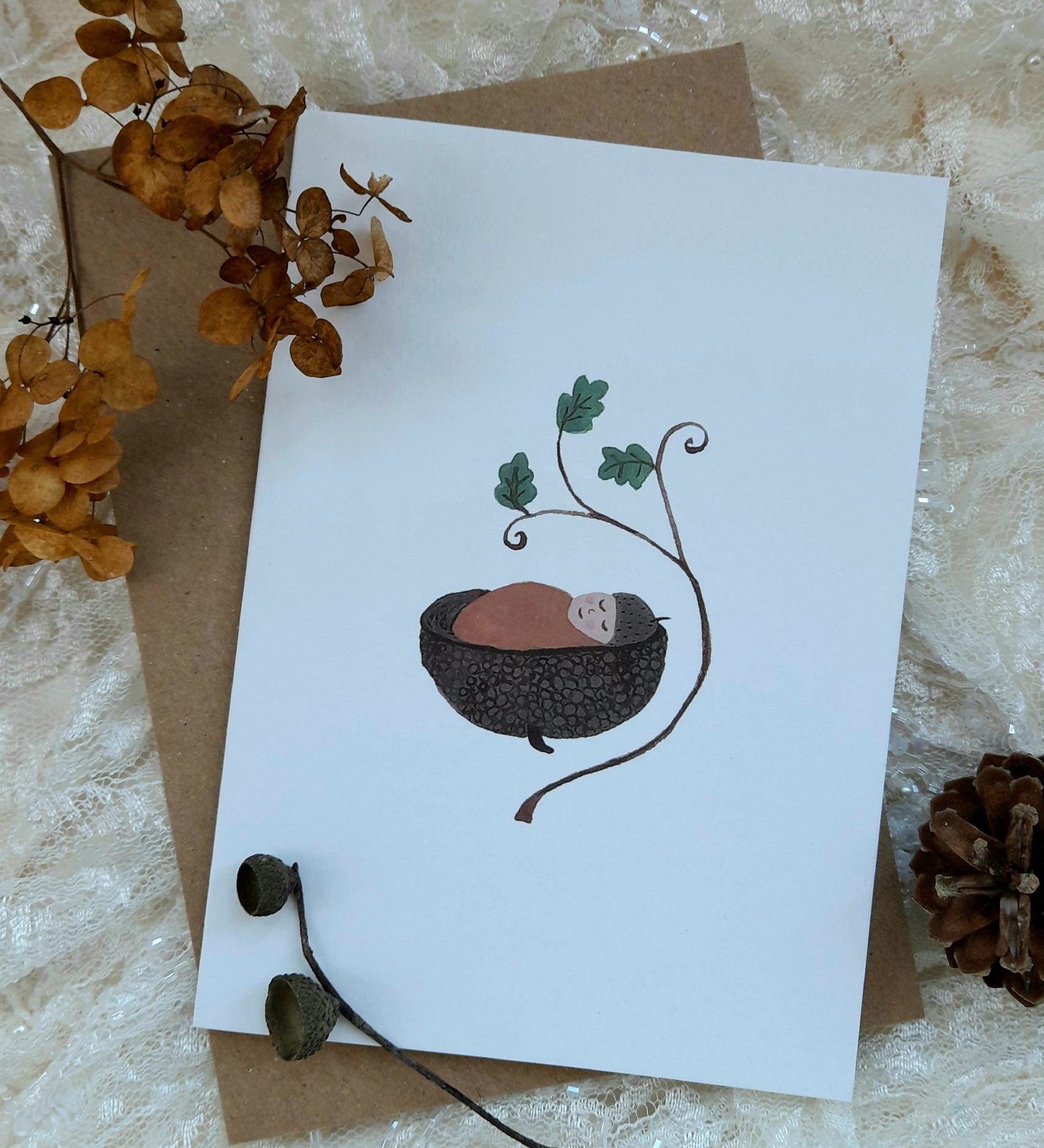 Acorn baby greetings card from Lydiamae showing a illustration of a baby inside a acorn shell under a stick in a soft drawing style. Card is on top of a brown envelope