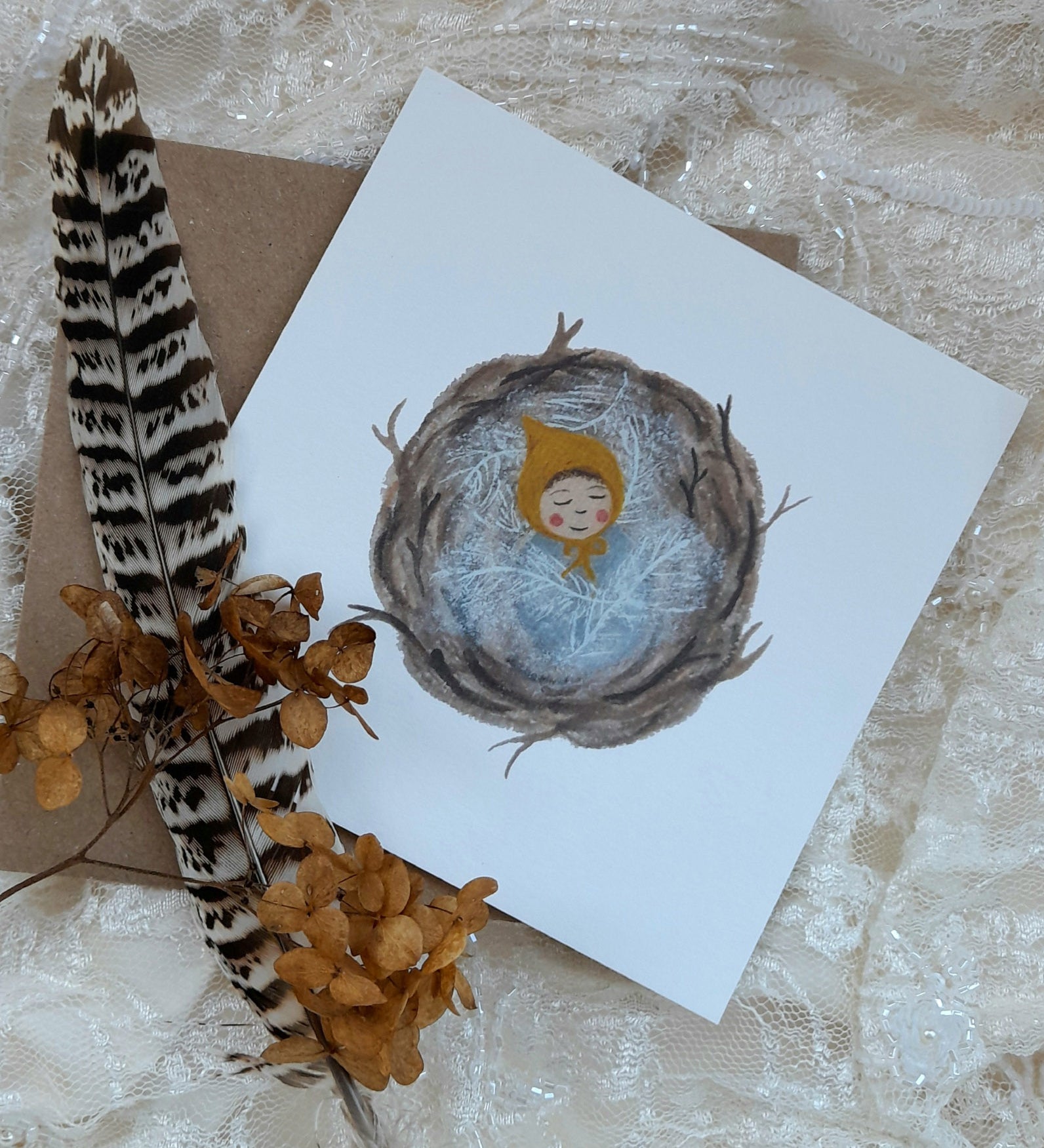 A baby nest greetings card from Lydiamae showing a illustration of a baby inside a birds nest in a soft drawing style. Card lying next to a feather