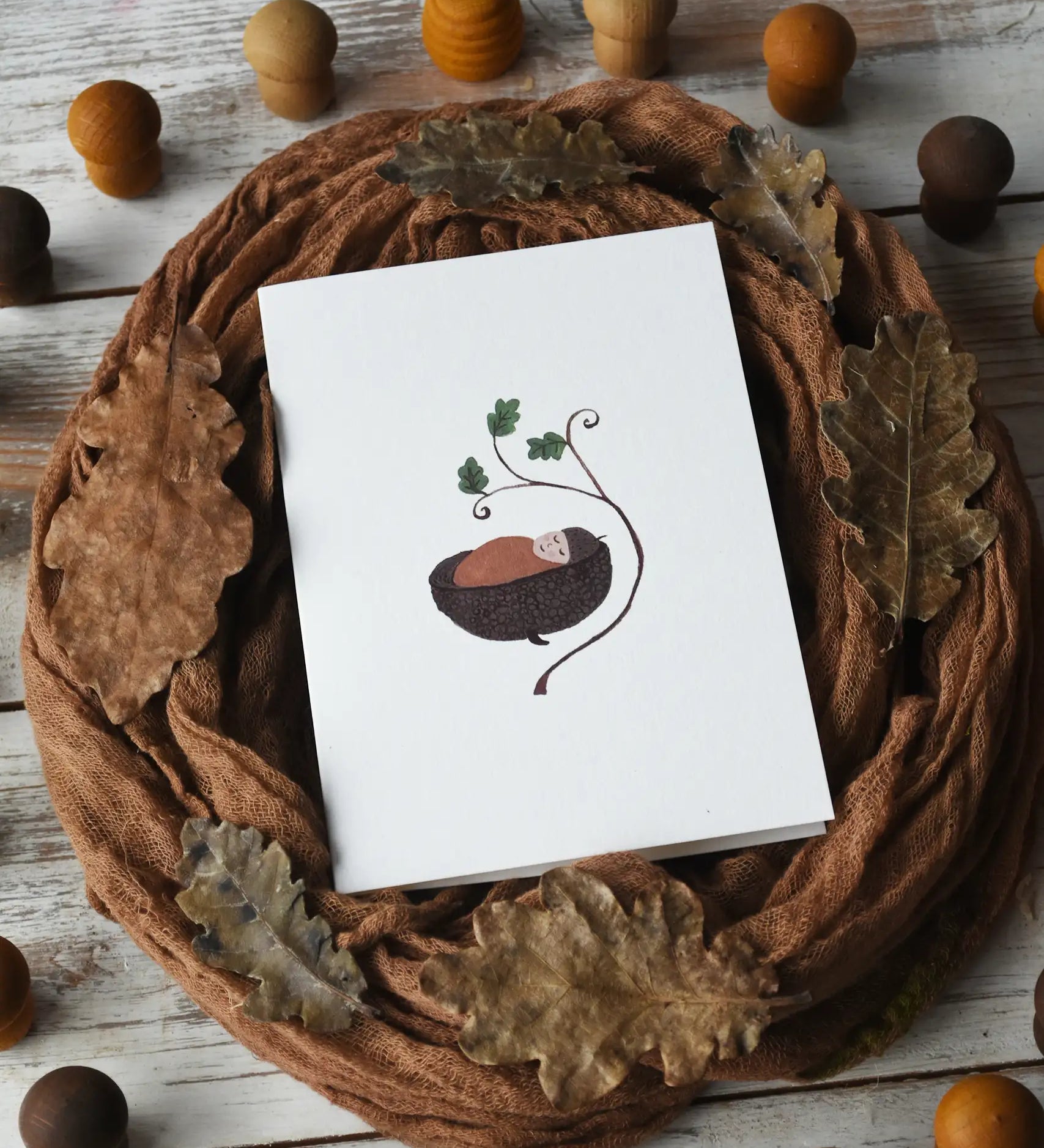 Close up of the LydiaMae acorn baby greetings card placed in the middle of a flatlay surrounded by a brown Ostheimer scarf covered in dried oak leaves and Grapat mandala pieces.