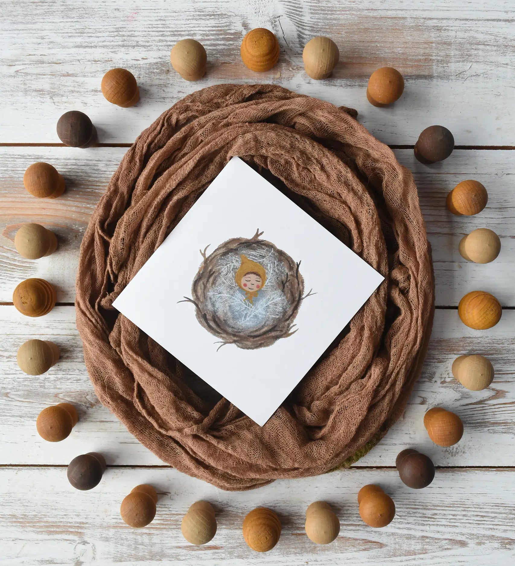 LydiaMae baby nest greetings card placed in the centre of a flatlay with a nest made with a brown Ostheimer scarf surrounded by brown. beige and yellow mandala pieces. 