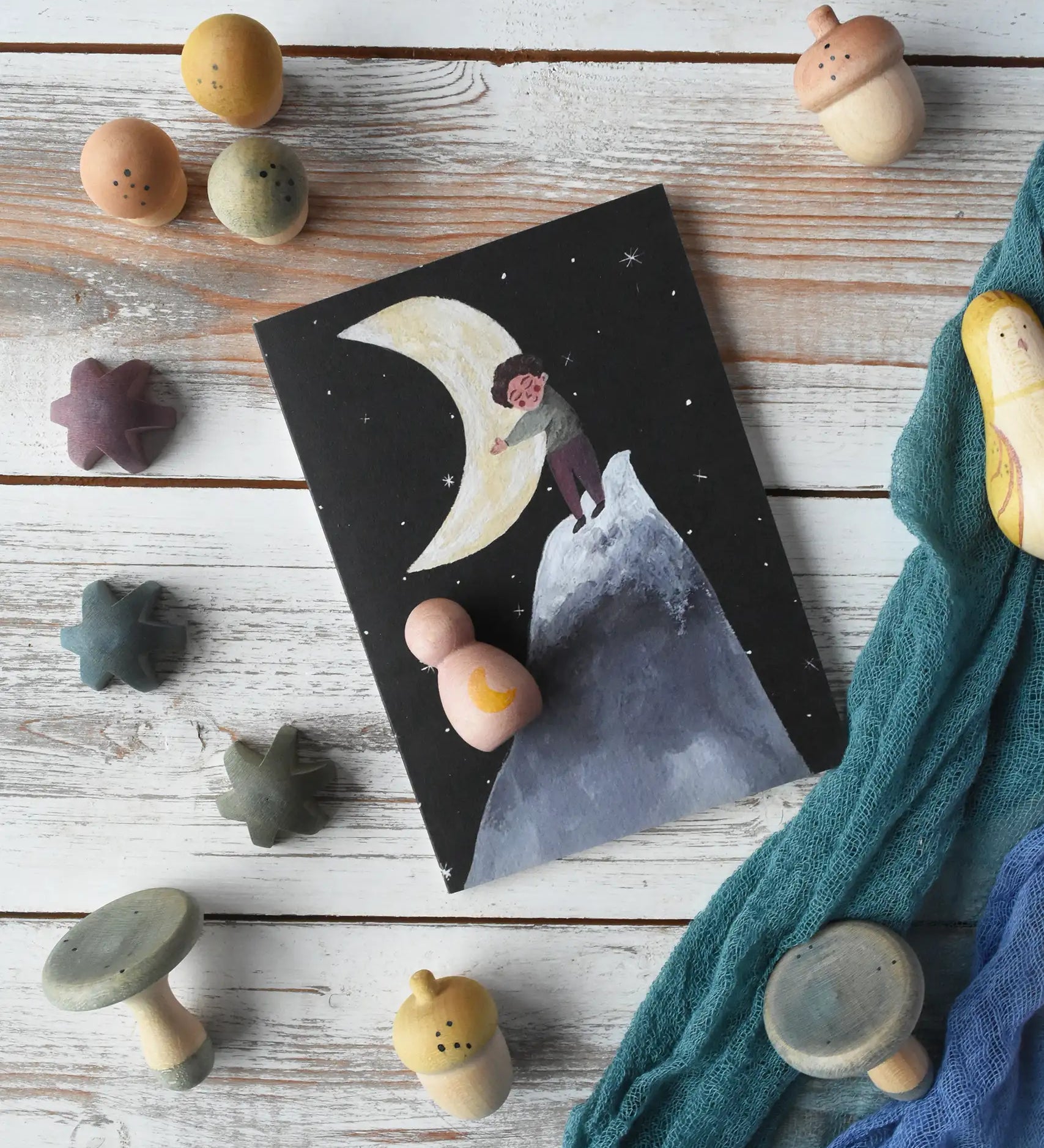  LydiaMae Hug The Moon Greetings Card placed on a white wooden surface in a flatlay with Ostheimer scarfs and wooden loose parts from the Grapat moonlight tale play set.