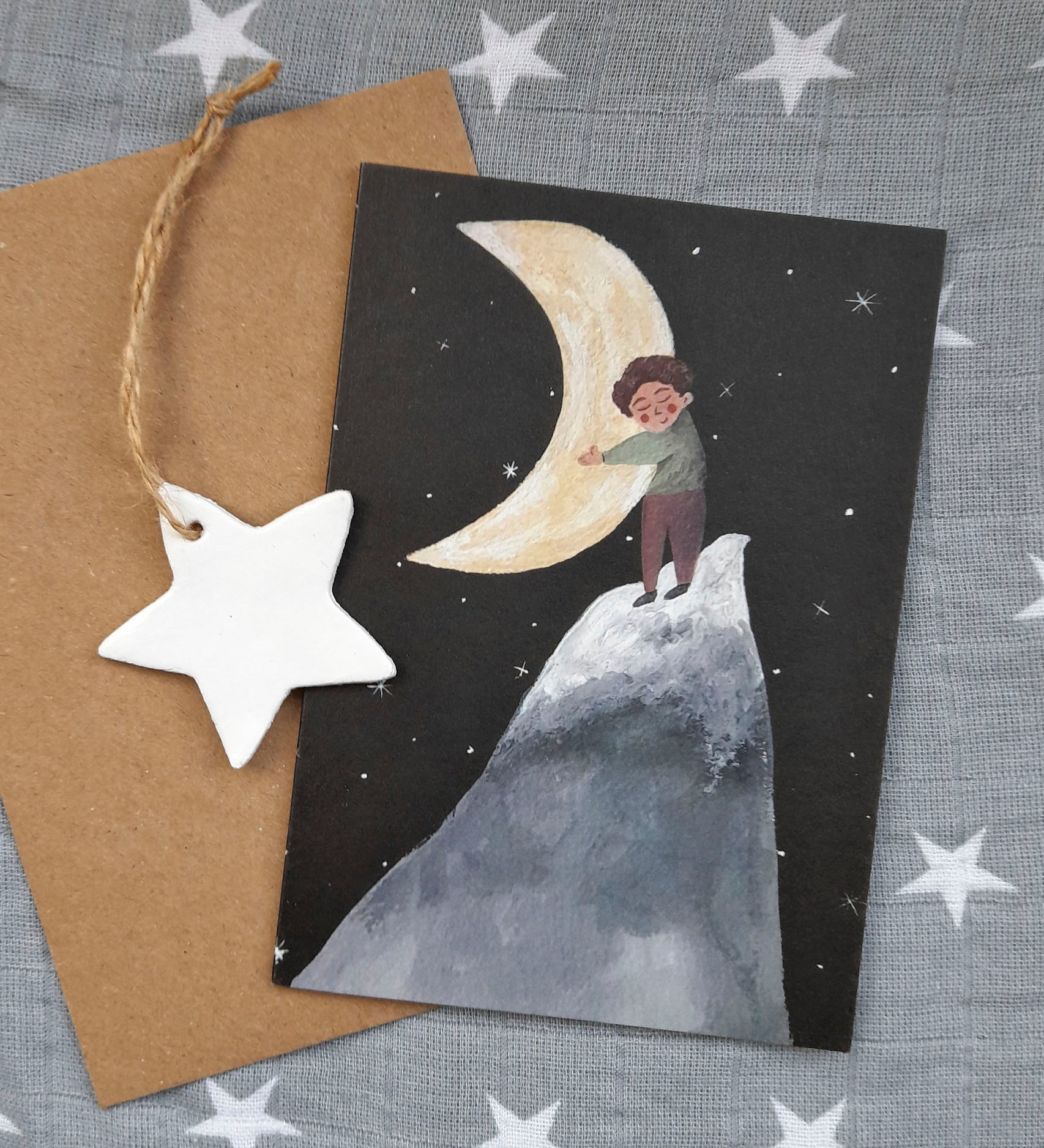 Hug the moon greetings card from Lydiamae showing a illustration of a child hugging the moon in a soft drawing style. Card is on top of a brown envelope