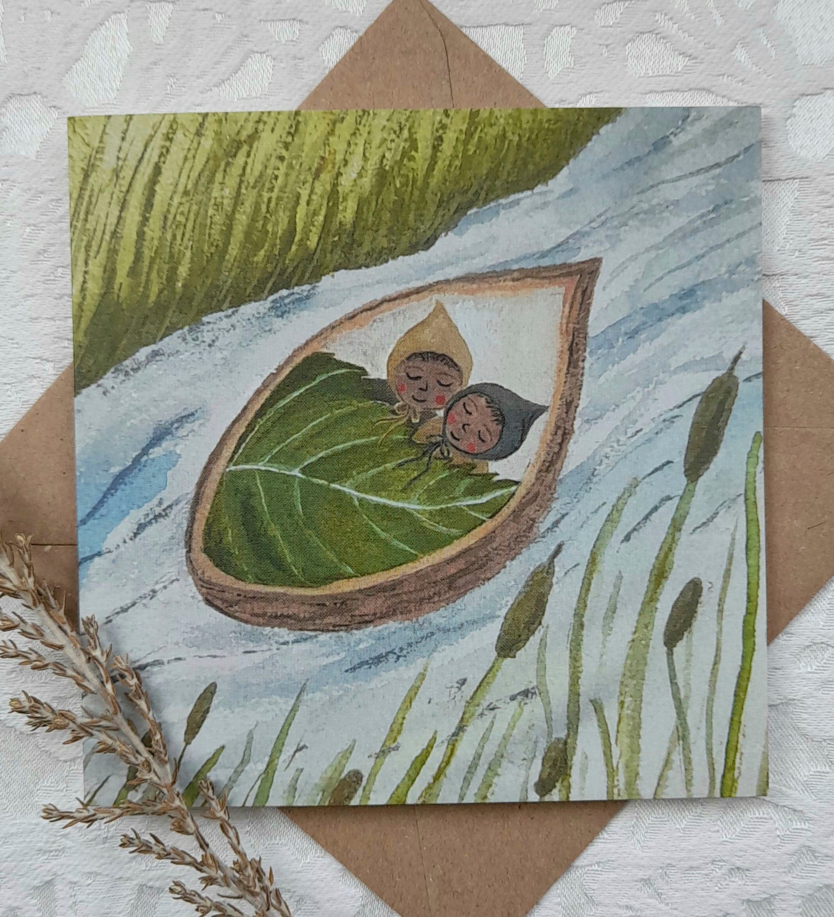A in a nutshell twins greetings card from Lydiamae showing a illustration of twins inside a shell of a nut floating down a river in a soft drawing style. Card is on top of a brown envelope