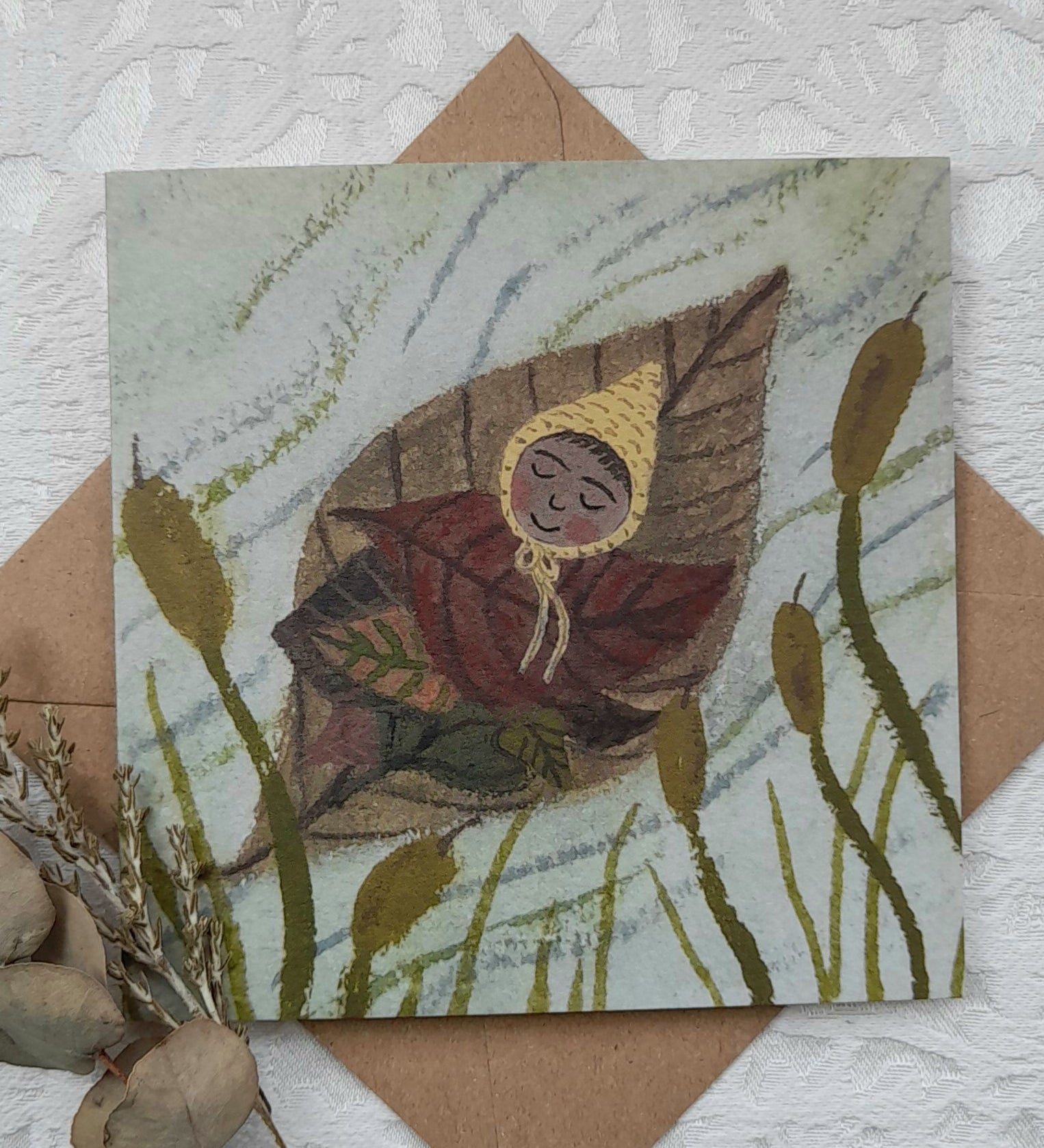 Leaf baby greetings card from Lydiamae showing a illustration of a baby on a leaf floating down a river in a soft drawing style. Card is on top of a brown envelope