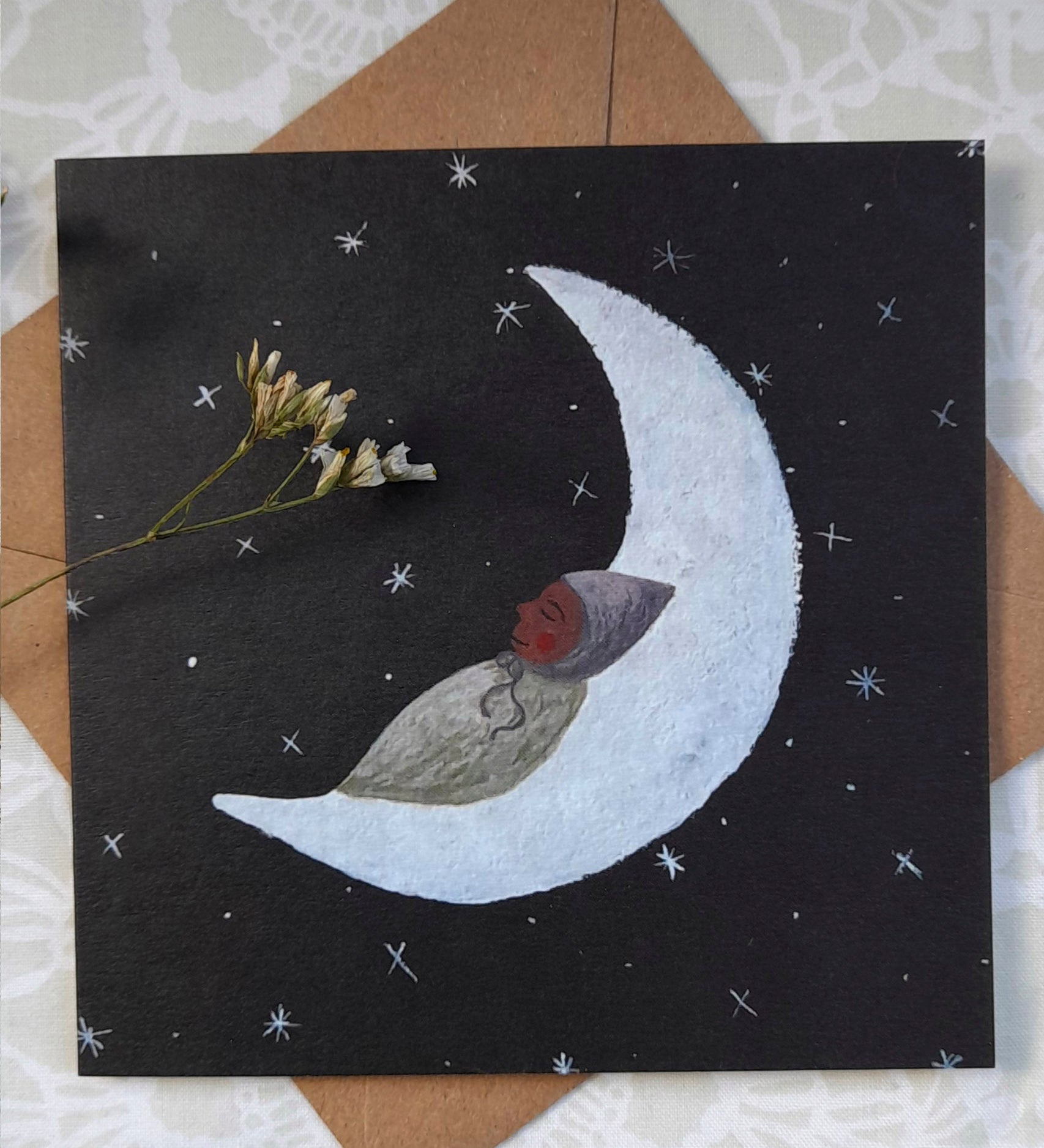 Moon baby greetings card from Lydiamae showing a drawing of a baby sleeping on the moon in a soft drawing style. Card is on top of a brown envelope