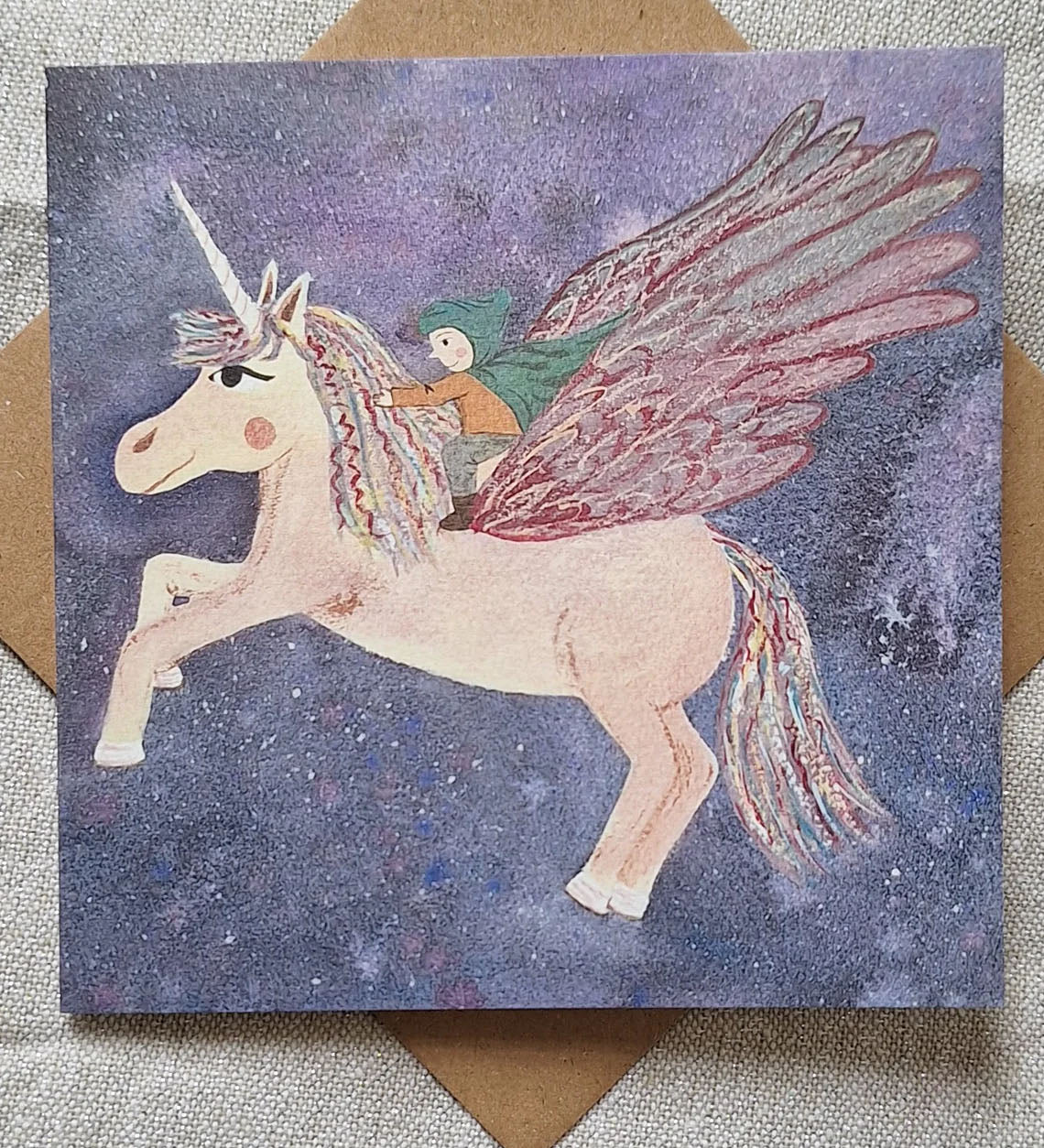 Pegasus greetings card from Lydiamae showing a drawing of a child riding a flying pegasus in a soft drawing style. Card is on top of a brown envelope
