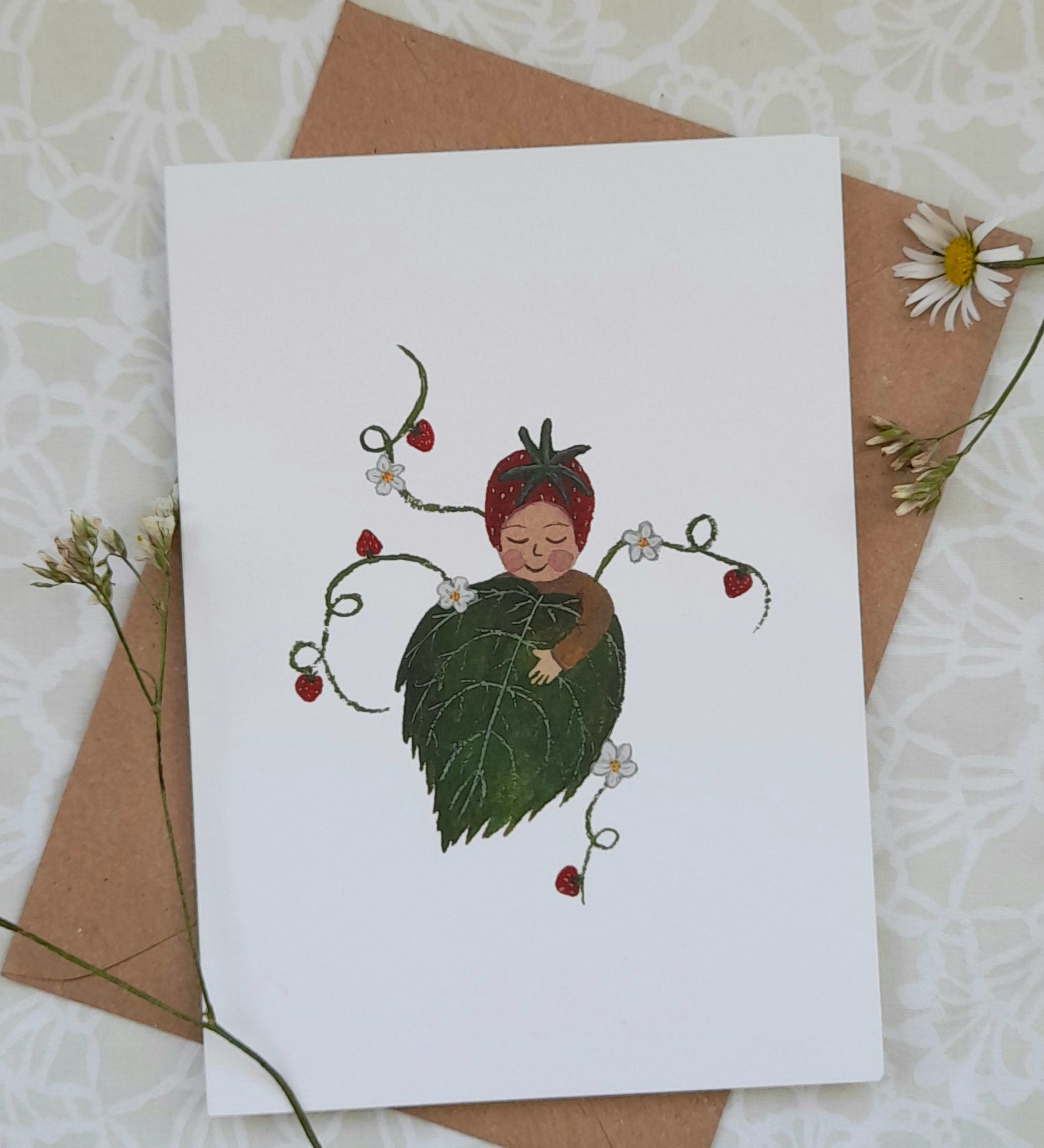 Strawberry baby greetings card from Lydiamae showing a drawing of a baby with a strawberry on the head in a soft drawing style. Card is on top of a brown envelope