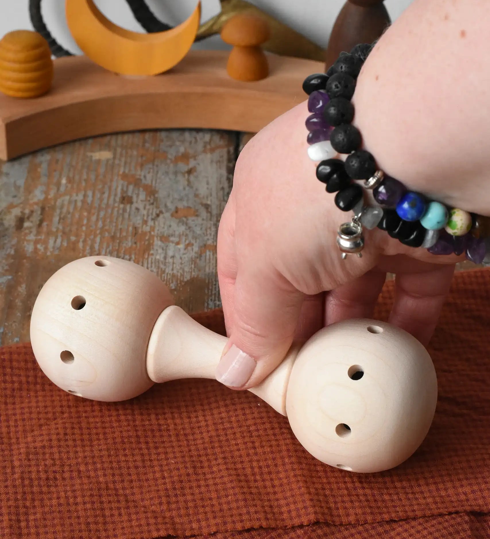 Mader Kreiselmanufaktur traditional wooden baby rattle placed on a cloth on a wooden surface in the Babipur playroom. An adult's hand can be seen picking the toy rattle up.