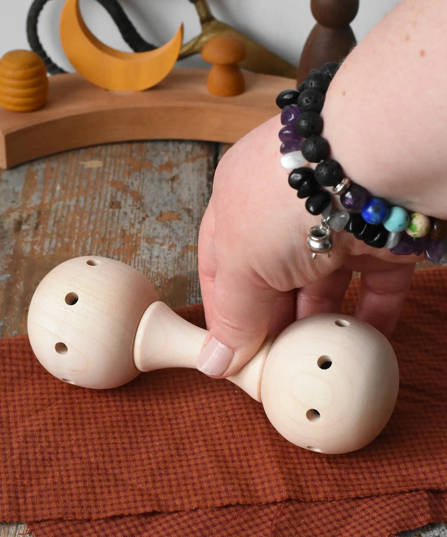 Mader Kreiselmanufaktur traditional wooden baby rattle placed on a cloth on a wooden surface in the Babipur playroom. An adult's hand can be seen picking the toy rattle up.