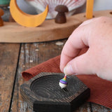 Mader hexagon 8cm spinning top plates in black oak with a tiny rainbow spinning top being placed on it.