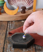 Mader hexagon 8cm spinning top plates in black oak with a tiny rainbow spinning top being placed on it.