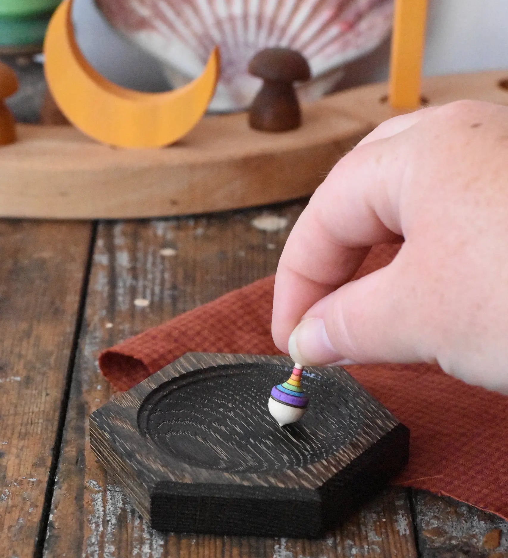 Mader hexagon 8cm spinning top plates in black oak with a tiny rainbow spinning top being placed on it.