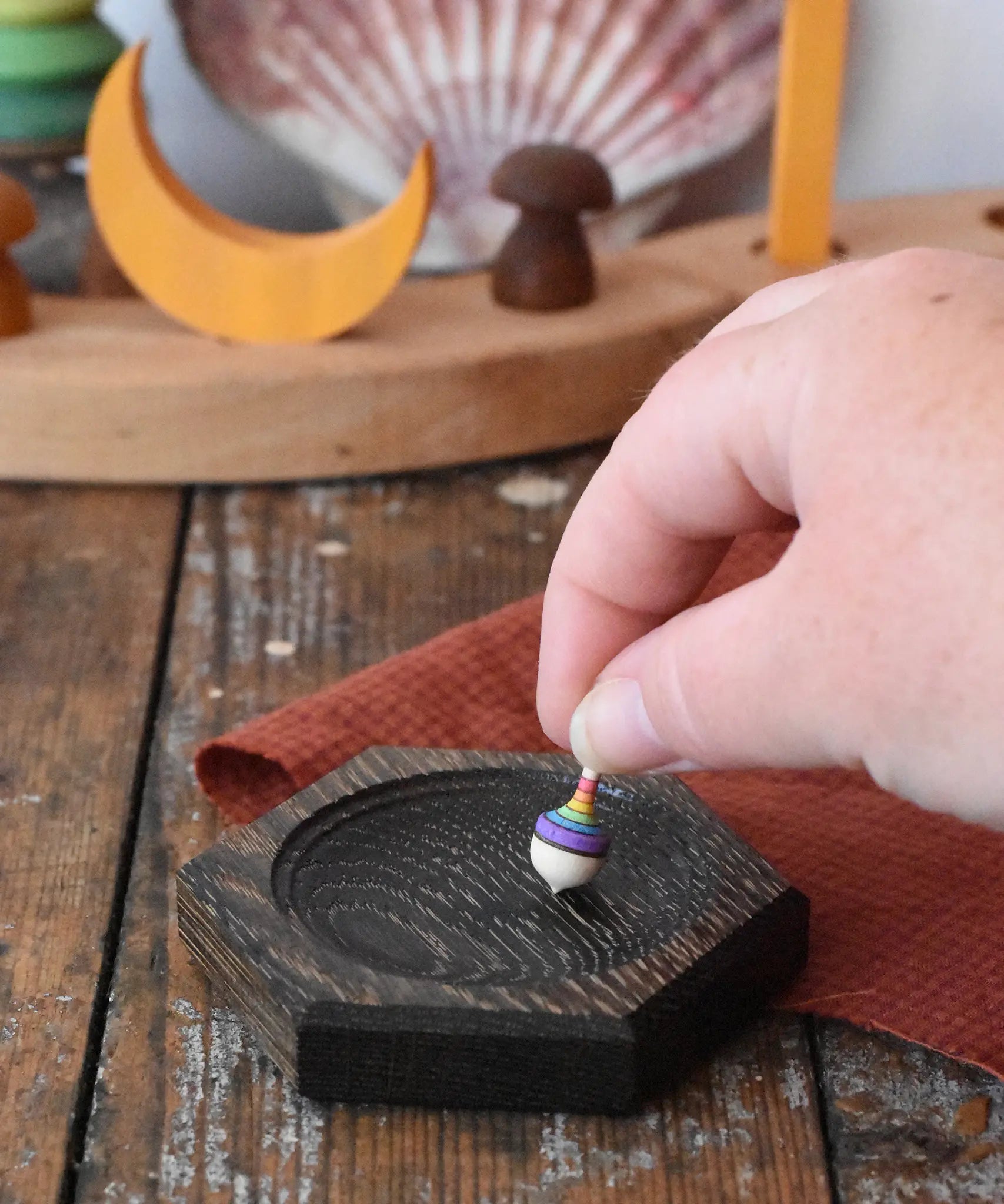 Mader hexagon 8cm spinning top plates in black oak with a tiny rainbow spinning top being placed on it.