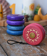 Mader burlesk wooden yoyo in pink placed on a wooden surface in the Babipur playroom. Other colours of the yoyo can be seen in the background.