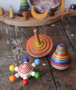 Mader carousel wooden baby grapsing toy with rainbow beads, orange first spinning top and striped roly poly pear toy placed on a wooden surface in front of a decorated Grimm's celebration ring.