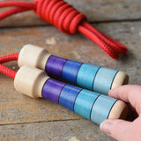 Mader Kreiselmanufaktur Children's Skipping Rope with Purple Handles placed on a wooden surface in the Babipur playroom. An adult's hand can be seen reaching for it.