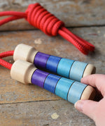 Mader Kreiselmanufaktur Children's Skipping Rope with Purple Handles placed on a wooden surface in the Babipur playroom. An adult's hand can be seen reaching for it.