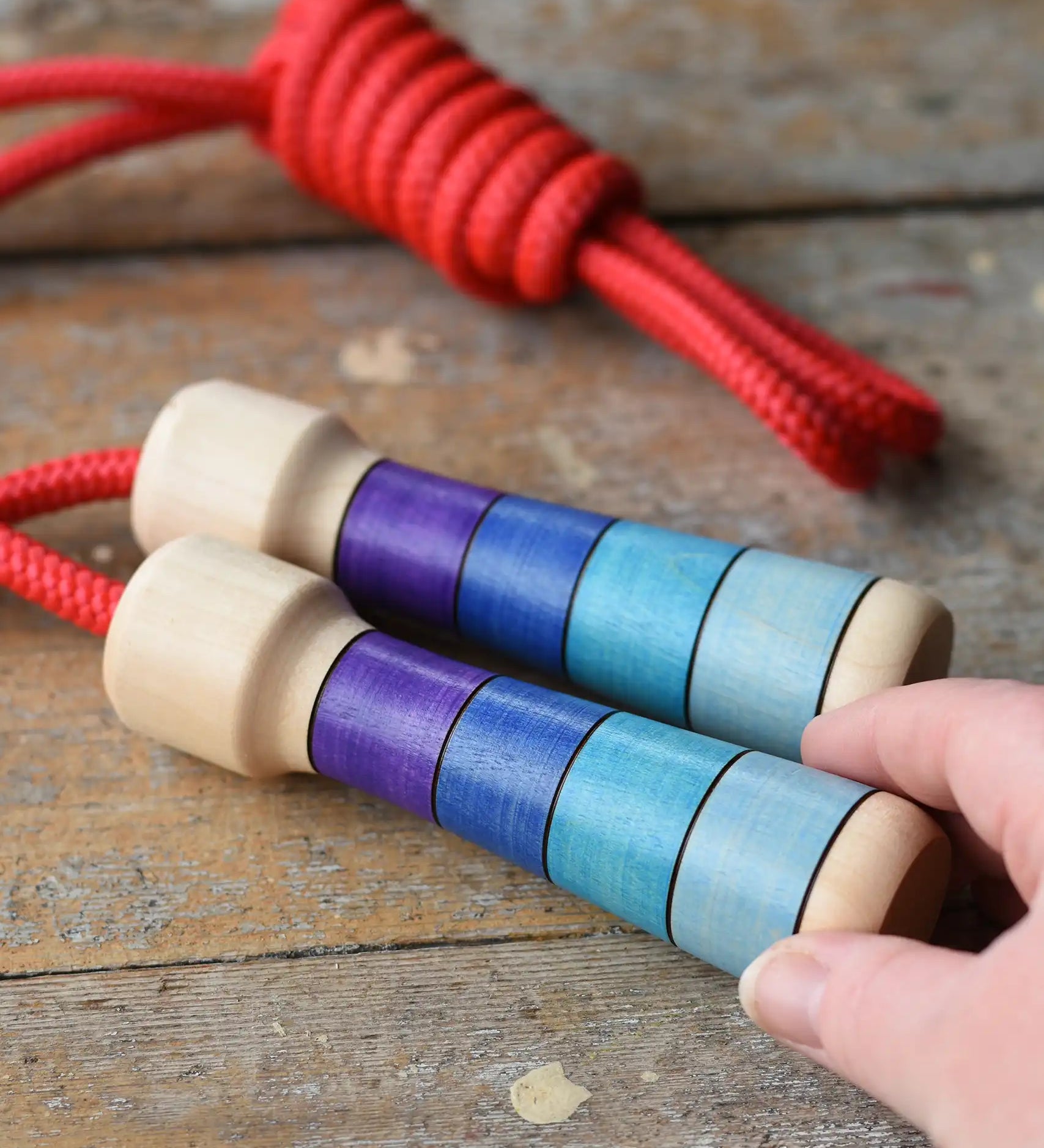 Mader Kreiselmanufaktur Children's Skipping Rope with Purple Handles placed on a wooden surface in the Babipur playroom. An adult's hand can be seen reaching for it.