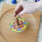 An adults hand placing the Mader So and So spinner on a wooden spinning plate. The spinner has one of the colourful geo print discs placed on it. 