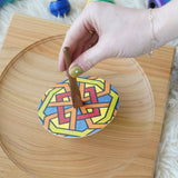 An adults hand placing the Mader So and So spinner on a wooden spinning plate. The spinner has one of the colourful geo print discs placed on it. 