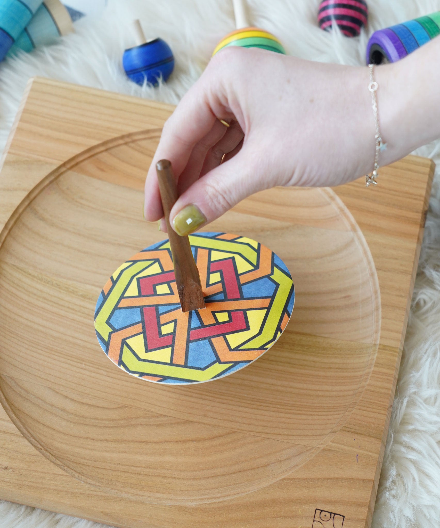 An adults hand placing the Mader So and So spinner on a wooden spinning plate. The spinner has one of the colourful geo print discs placed on it. 