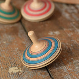 Mader Kreiselmanufaktur fado maple wooden finger spinning top placed on a wooden surface in the Babipur playroom. Other colour variants can be seen in the background. 
