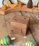 Mader Kreiselmanufaktur forest music box placed on a wooden desk in the Babipur playroom. A wooden wind up musical box with two slots for the green tree shaped spinners on the top. The two trees have been placed besides the box.