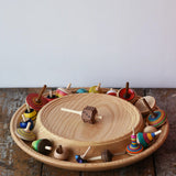 Mader Kreiselmanufaktur 40cm Rondell Ash Wood Spinning Plate placed on a wooden surface in the Babipur playroom. Dice spinning top placed on the top with other spinning tops filling bottom part of the plate. 