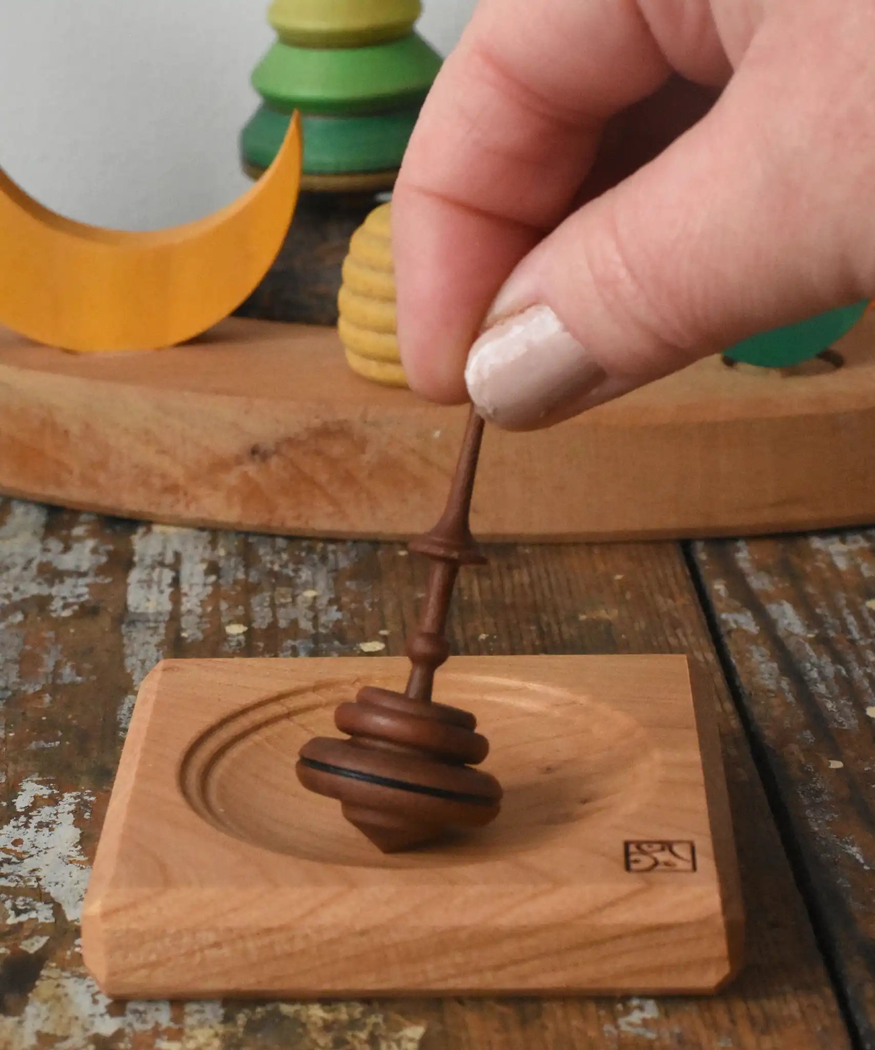 Mader Helene's captive rings small plum spinning top with mini board placed on a wooden surface in the Babipur playroom. An adult's hand can be seen reaching for the spinning top.