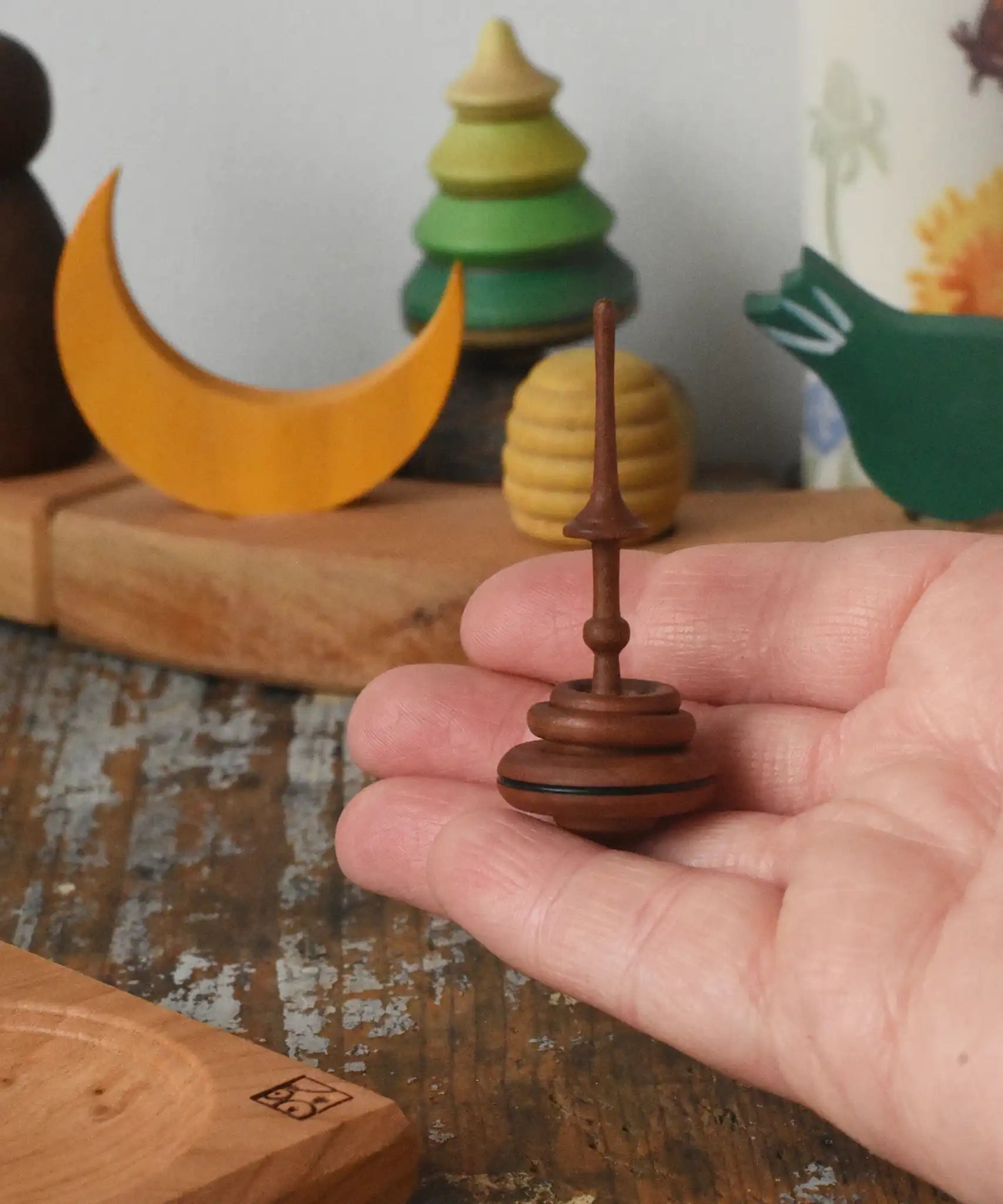 Mader Helene's captive rings small plum spinning top shown in an adult's hand with mini board in the background placed on a wooden surface in the Babipur playroom.