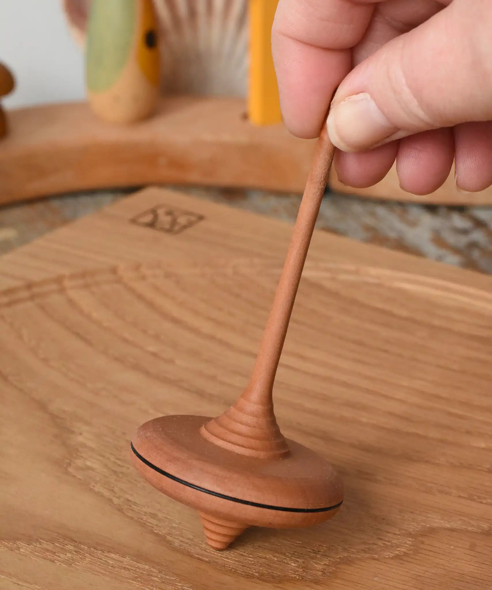 Mader ibis spinning top placed on a wooden desk in the Babipur playroom. The top is held upright by and adult's hand.