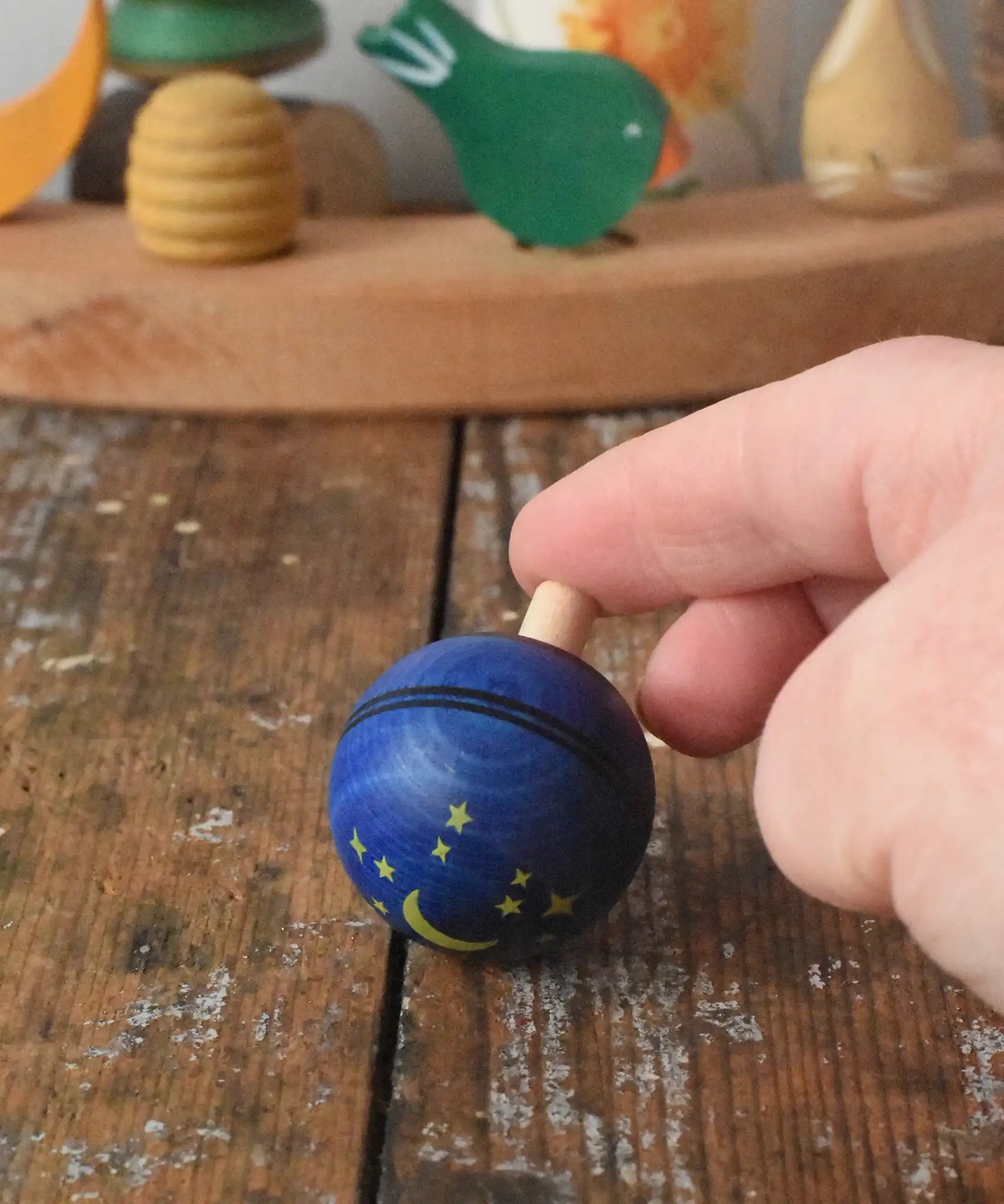 Mader Luna turn over spinning top shown on a wooden desk in the Babipur playroom. An adult's hand is holding it on a slant.