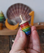 Mader handcrafted wooden rainbow mini thunderbolt spinning top in an adult's hand with a decorated Grimm's celebration ring in the background.